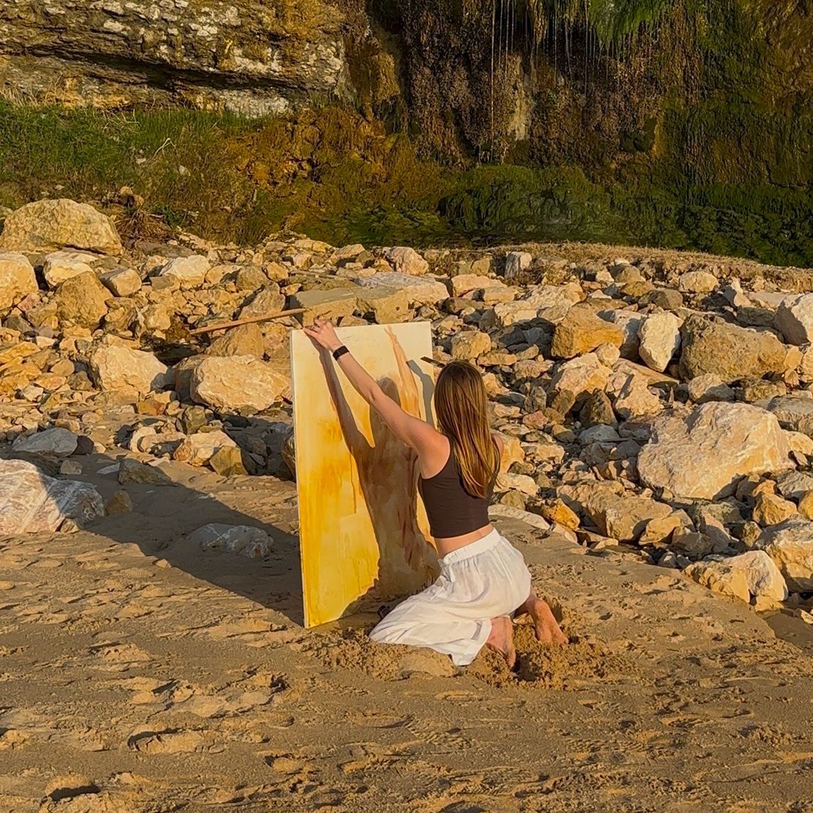 The artist kneeling on the beach, painting a canvas propped in the sand